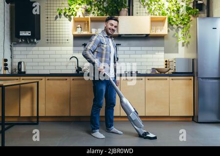 Porträt eines Mannes zu Hause, der Hausarbeit leistet, den Boden in der Küche zu Hause saugt, die Kamera anschaut und lächelt Stockfoto