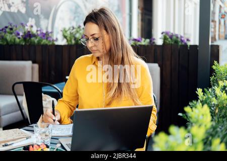 Zeitmanagement für Freiberufler. Freiberufliche Mitarbeiterin schreibt in einem Notebook und arbeitet mit einem Laptop Stockfoto