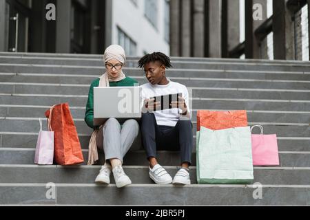 Outdoor in voller Länge Fashion Portrait von zwei jungen multirassischen Damen in stilvoller Kleidung sitzen auf den Stufen eines großen Einkaufszentrums und bestellen Kleidung online farbige Einkaufstaschen liegen neben ihnen Stockfoto