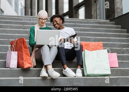 Outdoor in voller Länge Fashion Portrait von zwei jungen multirassischen Damen in stilvoller Kleidung sitzen auf den Stufen eines großen Einkaufszentrums und bestellen Kleidung online farbige Einkaufstaschen liegen neben ihnen Stockfoto