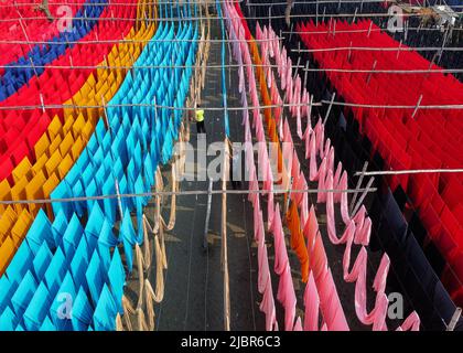 8. Juni 2022, Narayanganj, Dhaka, Bangladesch: Arbeiter hängen Tausende von Metern verschiedenfarbiger Stoffe zum Trocknen auf, damit sie zu T-Shirts und Westen verarbeitet werden können. Eisendrähte werden zwischen Bambusrahmen verwendet, um riesige Waschlinien für den letzten Teil des Sterbeprozesses zu schaffen, während die Stoffe in Narayanganj, Bangladesch, in der Sonne getrocknet werden. Helle Stränge aus blau, rosa, orange, rot, lila, Gelbe, magentafarbene und grün gefärbte Tücher hängen über dem grasbewachsenen Feld in einem blendenden Netzwerk von ineinanderschachernden Farben. Nach fünf bis sechs Stunden in der Sonne wird der Stoff für die Herstellung von Kleidern verwendet.Dies ist das Finale Stockfoto