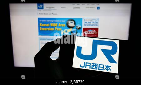 Person mit Mobiltelefon und Logo der japanischen Firma West Japan Railway (JR) auf dem Bildschirm vor der Business-Webseite. Konzentrieren Sie sich auf die Telefonanzeige. Stockfoto