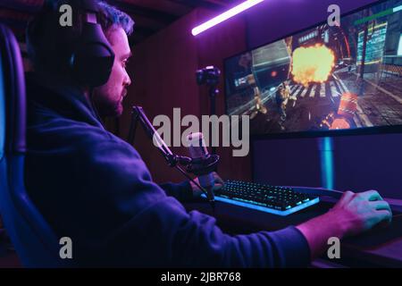 Seitenansicht von Pro Streamer spielen Videospiel Wettbewerb Verwenden Sie professionelle Gaming-Setup. Spieler, der spät in der Nacht zu Hause auf einem Spielstuhl sitzt und professionelle Kopfhörer und Mikrofon verwendet. Cyber-Sport Stockfoto