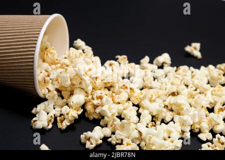 Popcorn wird aus einem braunen Papierbecher auf schwarzem Hintergrund in Nahaufnahme ausgestreut Stockfoto