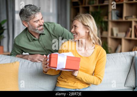 Liebevoller Mann mittleren Alters, der seine Frau zu Hause mit einem Geschenk überraschte Stockfoto