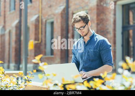 Junger Geschäftsmann mit Brille in der Nähe des Büros, der an einem Laptop arbeitet Stockfoto