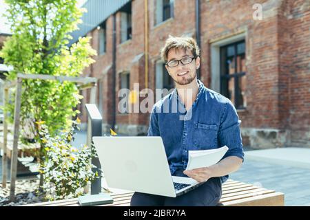 Porträt eines jungen Lehrers außerhalb des Universitätscampus, der mit einem Laptop auf einer Bank sitzt und lächelt Stockfoto