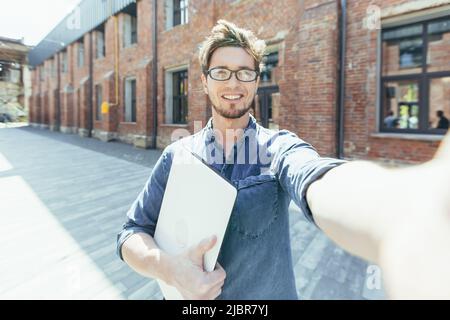 Fröhlicher junger Student, der lächelnd und mit der Hand winkend auf die Smartphone-Kamera schaut, mit Freunden im Videogespräch spricht, Mann draußen auf dem Campus Stockfoto