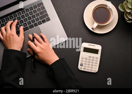 Geschäftsfrau oder Buchhalterin, die an ihrem Schreibtisch arbeitet und auf einem Notebook-Laptop tippt. Laptop, Rechner und Kaffeetasse auf schwarzem Hintergrund Stockfoto
