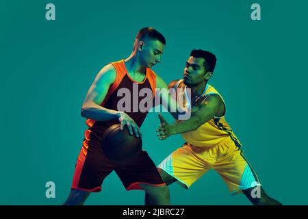 Porträt von zwei jungen Männern, Basketballspieler im Uniformtraining isoliert auf blauem Hintergrund in Neonlicht. Position des Verteidigers. Stockfoto