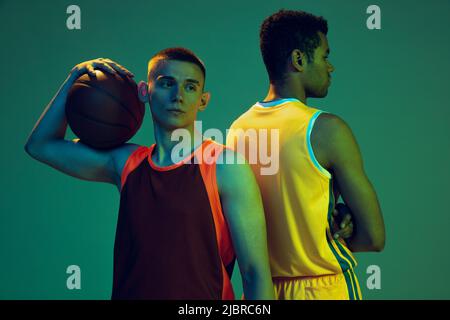 Porträt von zwei jungen Männern, professionellen Basketballspielern, die sich in Neonlicht isoliert auf blauem Studiohintergrund auflehnen. Team Stockfoto