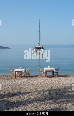 Bodrum, Mugla, Türkei. April 22. 2022 Restauranttische am Strand mit einem Katamaran im Hafen von Bodrum, einem beliebten Ziel für Segeln und Kreuzfahrt Stockfoto