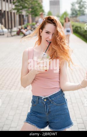 Junge schöne rothaarige Frau mit Zahnspange Getränke kühlen Cocktail im Freien im Sommer. Porträt eines lächelnden Mädchens mit Sommersprossen Stockfoto