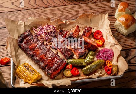 Gegrillte Schweineribs, serviert mit gegrilltem Mais, Salat, bbq-Sauce, Salz, Pfeffer und Gurke auf Pergamentpapier auf einem Holztisch. Draufsicht Stockfoto