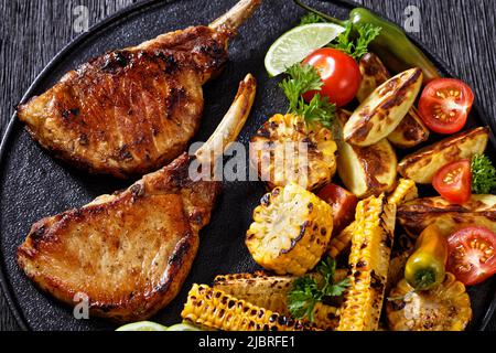 Gebratene Tomahawk-Schweinekoteletts, Einbeinsteak mit gegrillten Maisscheiben und Maisrippen, Keile-Kartoffeln, frische Tomaten auf schwarzem Teller auf dunklem Holztisch, Stockfoto