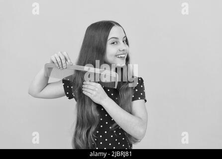 Fröhliches Kind bürstet lange Haare mit Kamm auf gelbem Hintergrund, Friseur Stockfoto