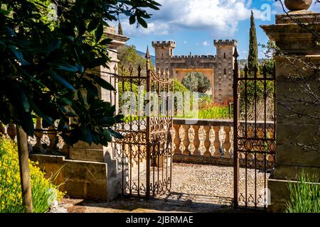 Ein rostiger, reich verzierter Eisenzaun vor einer Steinmadengeländer führt in einen Garten mit grünen Pflanzen, der den beeindruckenden Eingang von Raixa umgibt. Stockfoto