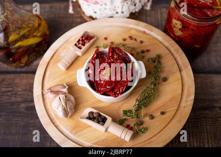 Sonnengetrocknete Tomaten in Olivenöl in einer weißen Schüssel mit Pfeffer, Knoblauch und Thymian auf einem Holzbrett. Hausmannskost, rustikaler Stil. Selektiver Fokus, Nahaufnahme Stockfoto