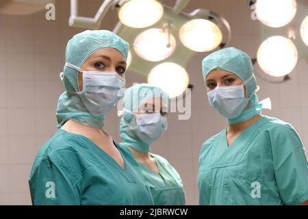 Drei Krankenschwestern werden in einem Operationssaal gesehen. Sie sind als OP-Krankenschwestern mit Gesichtsmasken und steriler OP-Kleidung gekleidet Stockfoto