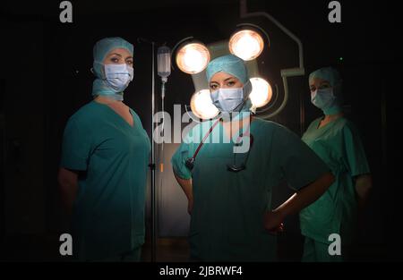 Drei Krankenschwestern werden in einem Operationssaal gesehen. Sie sind als OP-Krankenschwestern mit Gesichtsmasken und steriler OP-Kleidung gekleidet Stockfoto