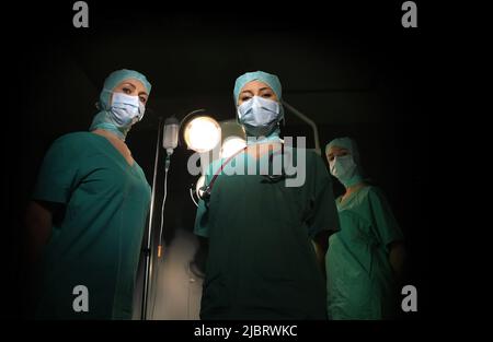 Drei Krankenschwestern werden in einem Operationssaal gesehen. Sie sind als OP-Krankenschwestern mit Gesichtsmasken und steriler OP-Kleidung gekleidet Stockfoto