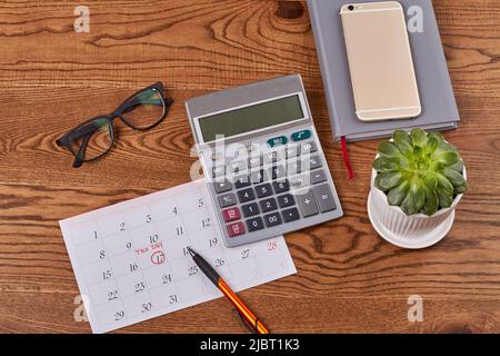 Business-Accessoires auf braunem Holzschreibtisch. Kalender mit Steuertag mit Rechner und Brille. Grüne Pflanze im Topf. Stockfoto