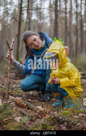 Mutter und Kind, die nach dem Regen in Regenmänteln gemeinsam im Wald spazieren gehen, Pilze auf einem umgestürzten Baum betrachten und reden Stockfoto