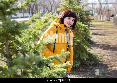 Eine brünette Frau in einer gelben Jacke guckert hinter einem Baum auf einem Spaziergang hervor. Stockfoto