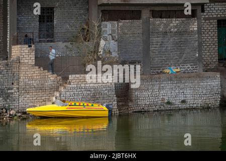 Helles gelbes Schnellboot, das an einer Blockmauer und einem halb gebauten Haus am Ufer des Nils, Ägypten, festgemacht ist Stockfoto