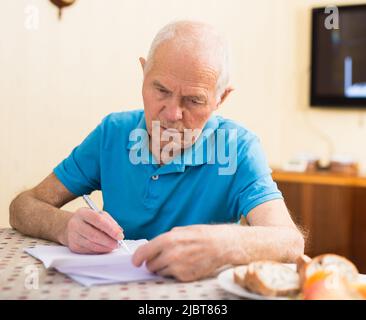 Fokussierter älterer Mann, der zu Hause sitzt und Papiere liest und unterschreibt Stockfoto