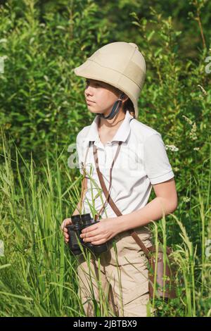 Das Mädchen mit dem Pithhelm steht im Gras und hält ein Fernglas und beobachtet die Natur des Sommers. Discovery and Adventures Konzept Stockfoto