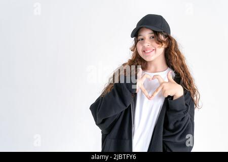 Stilvolles Teenager-Mädchen mit langen lockigen Haaren in einem schwarzen Hoodie und einer schwarzen Mütze zeigt mit ihren Fingern eine Herzform und lächelt auf weißem Hintergrund. Platz Stockfoto