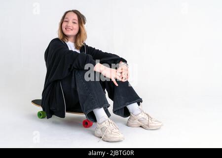 Ein stylisches Teenager-Mädchen mit Bob-Frisur in einem schwarzen Hoodie und schwarzer Jeans sitzt auf einem Longboard mit Kopfhörern und lächelt auf weißem Hintergrund. Spac Stockfoto