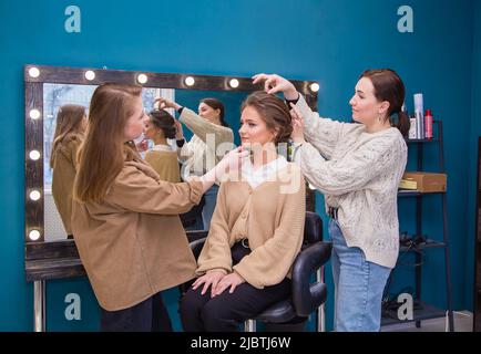 Zwei Meister stylen das Haar einwandfrei und bilden das Mädchen im Salon. Make-up-Künstler und Friseur schaffen ein Bild einer jungen Frau. Geschäftskonzept - Schönheitssalon, Gesichtshaut- und Haarpflege. Stockfoto