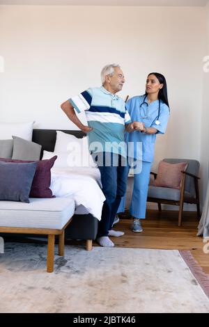 Biraziale Physiotherapeutin, die kaukasischen älteren Mann mit Rückenschmerzen beim Stehen zu Hause unterstützt Stockfoto