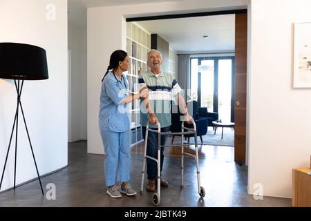 Biracial weibliche Physiotherapeutin Unterstützung kaukasischen älteren Mann beim Gehen mit Wanderer zu Hause Stockfoto