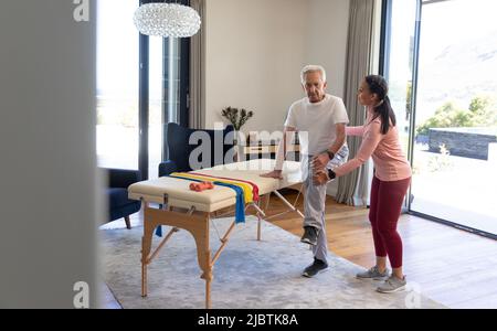 Biraziale Physiotherapeutin, die kaukasischen älteren Mann bei der Dehnung von Beinen an einem Tisch zu Hause unterstützt Stockfoto