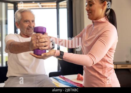 Lächelnde biracial weibliche Physiotherapeutin hilft kaukasischen älteren Mann beim Heben der Hantel zu Hause Stockfoto