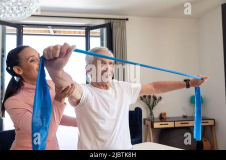 Biracial weibliche Physiotherapeutin Unterstützung kaukasischen älteren Mann Dehnung Widerstandsband zu Hause Stockfoto