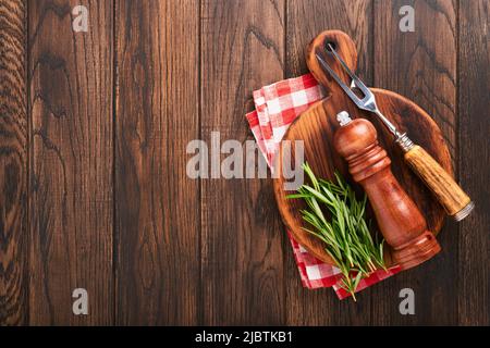 Hintergrund der Speisen. Grillgabel, Pfefferstreuer und Rosmarinzweig, rote Serviette auf Holzkochhintergrund. Hintergrund zum Kochen von Speisen. Zutaten zum Kochen Stockfoto