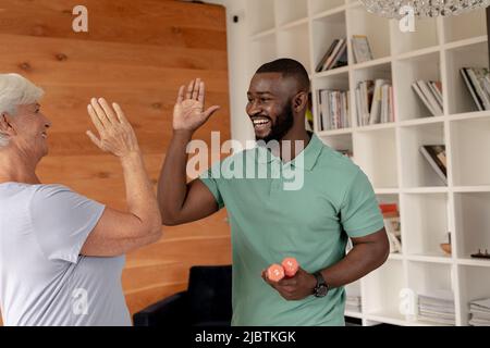 afroamerikanischer männlicher Physiotherapeut, der zu Hause Hanteln mit hohen Fiving kaukasischer älterer Frau hält Stockfoto