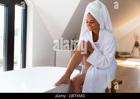 Kaukasische Frau in einem Bademantel, die Feuchtigkeitspflege auf ihr Bein auftragen und in der Nähe der Badewanne im Badezimmer sitzen Stockfoto