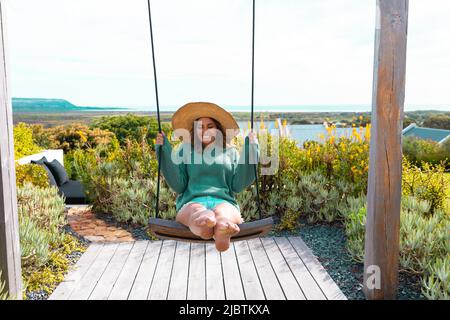 Kaukasische junge Frau, die einen Hut trägt und auf einer Schaukel im Freien schwingt Stockfoto