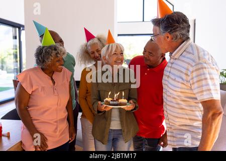 Multirassische Freunde, die aufgeregt ältere Frau betrachten, die im Altersheim Geburtstagskuchen hält Stockfoto