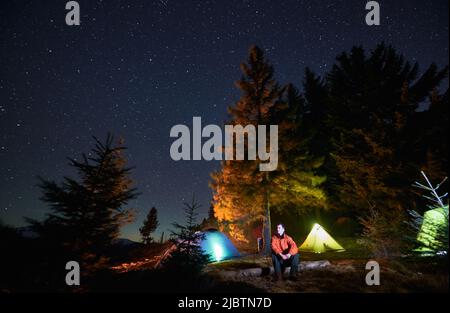 Ein männlicher Tourist, der auf Holzbalken in der Mitte der Lichtung sitzt und die wunderschöne Aussicht auf die Umgebung bei Nacht genießt. Im Hintergrund Wald, Campingplatz mit beleuchteten Zelten und Himmel mit Sternen übersät. Stockfoto