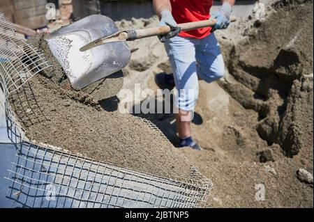 Nahaufnahme eines Mannes mit Schaufel in den Händen, der zementartige Bodenestriche mit Netz in einen Behälter warf. Männchen in Arbeitshandschuhen schaufeln Sand-Zement-Mischung im Freien auf der Baustelle. Stockfoto