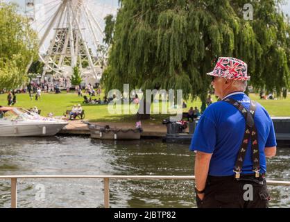 Ein Mann in einem Hut der Union Flag blickt über einen Fluss und sieht Menschen, die sich entspannen und die Sonne in einem Park genießen. Britisches Urlaubs-, Tourismus- oder Reisekonzept. Stockfoto