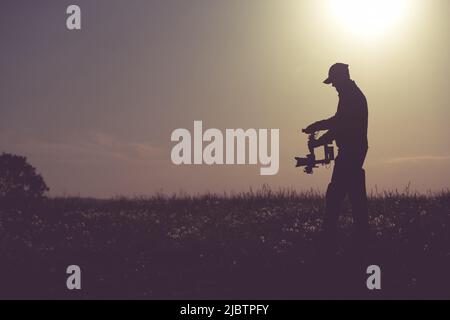 Silhouette eines männlichen Videografen in einem großen offenen Feld mit einer Kamera, die auf dem Gimbal-Stabilisator in seinen Händen platziert ist. Das Licht der aufgehenden Sonne im Bac Stockfoto