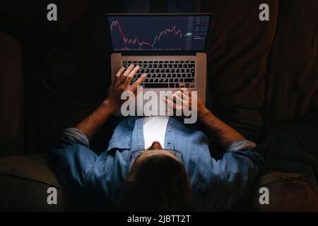 Der junge Geschäftsmann Trader-Analyst betrachtet den Laptop-Monitor auf dem Sofa, der Anlegerbroker analysiert Indizes, die Finanzchart, die Online-Investmentdaten auf dem Graphen des Kryptowährungsmarktes tradet. Draufsicht Stockfoto