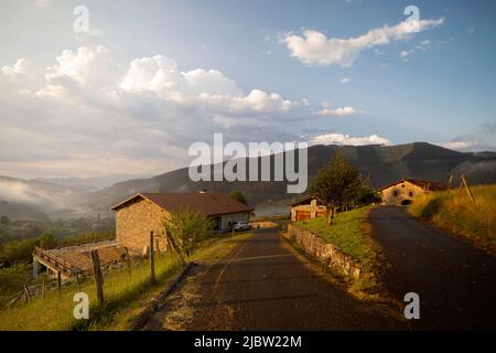 Bauernhäuser des Atxondo-Tals im Abendlicht mit dem Himmel mit flauschigen Wolken. Orange Töne dominieren im Bild. Naturbilder Stockfoto